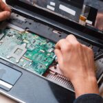 Kéz egy laptop belsejében, processzor cseréjéhez szükséges alkatrészekkel.