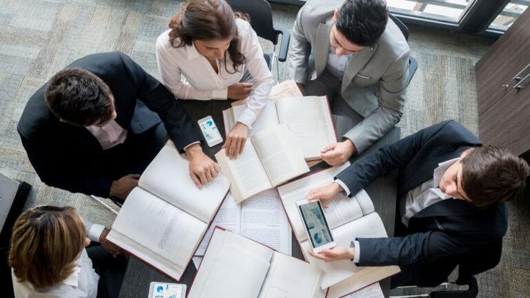 Öt üzletember egy asztal körül ülve, könyveket és táblagépet használva.